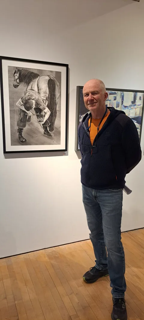 Kevin Swainson standing beside his charcoal drawing The Farrier II at an exhibition