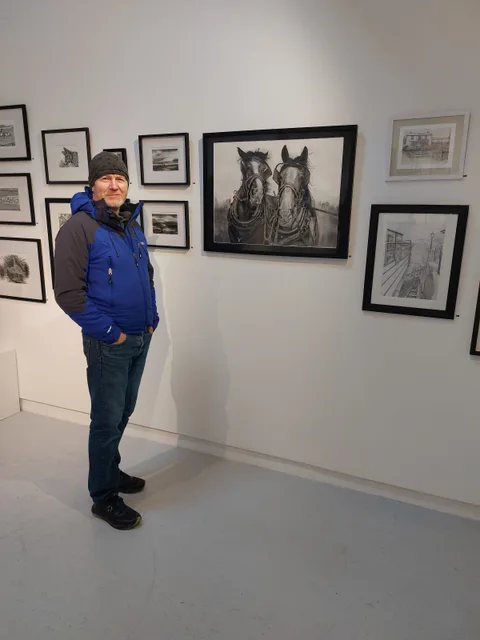 Kevin Swainson standing beside his charcoal drawing Working Horses at an exhibition